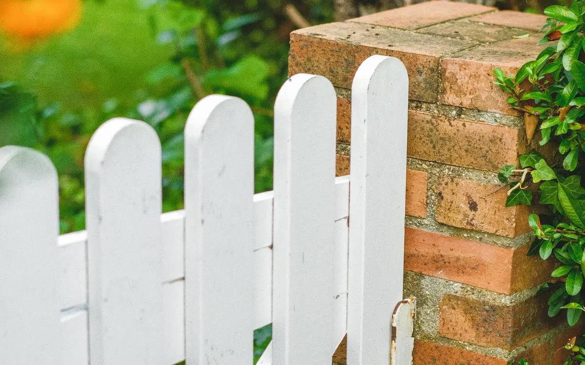 White wooden picket gate beside a brick pillar with greenery